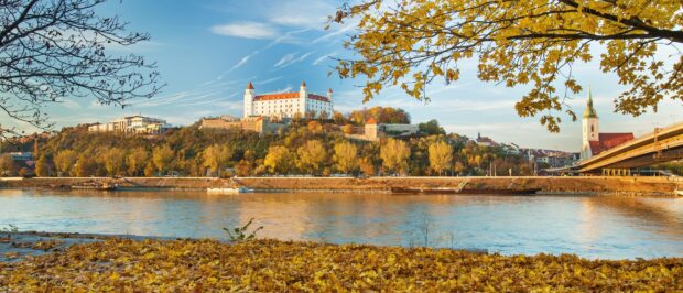 A scenic view of Bratislava castle surrounded by autumn trees and a river under a clear blue sky, 4K Desktop Wallpaper