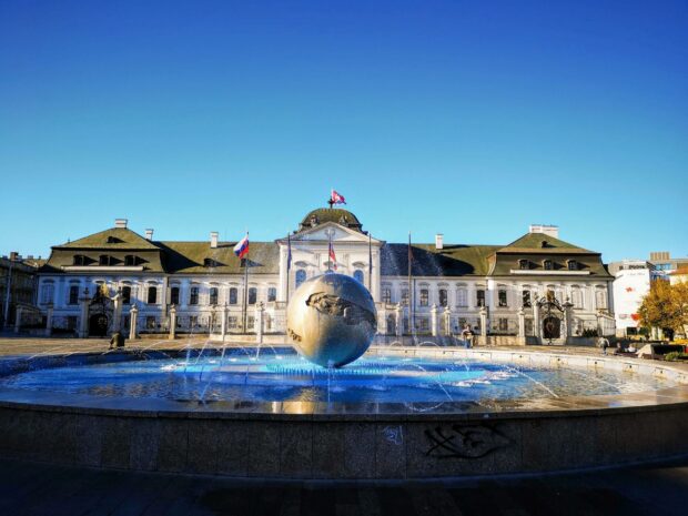 A large sphere fountain is in front of a historic building in Bratislava under a clear blue sky, 2K Desktop Wallpaper