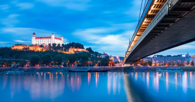 The Bratislava cityscape shows the illuminated castle and bridge over calm water during the evening, 2K Desktop Wallpaper