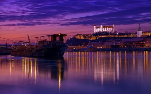 A beautiful view of Bratislava cityscape with illuminated castle and boats reflected in the river at sunset, HD Desktop Wallpaper