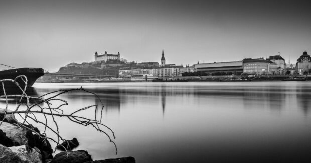 A peaceful view of Bratislava features historic buildings reflecting on the calm river waters, 2K Desktop Wallpaper