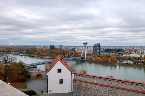 A scenic view of Bratislava with a river, bridge, and urban buildings under a cloudy sky, HD Desktop Wallpaper