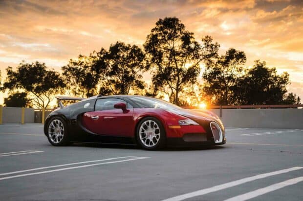 A red and black Bugatti Veyron is parked on an empty lot with a sunset sky and trees in the background, 2K Desktop Wallpaper