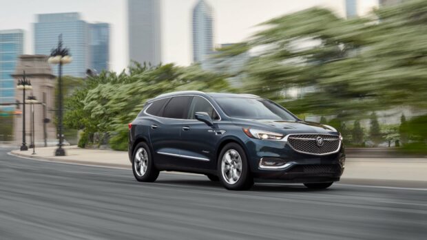 A Buick Enclave is driving smoothly on a city street with blurred buildings and greenery in the background, 4K Desktop Wallpaper