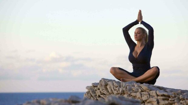 A woman meditating in a yoga pose on rocky cliffs near the ocean during sunrise, HD Desktop Wallpaper