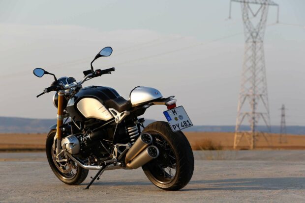 A BMW R Nine T motorcycle is parked on a wide open road with electrical towers in the background during sunset