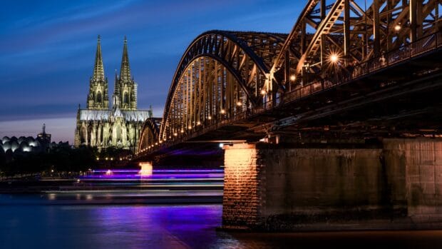 The Cologne cathedral towers stand illuminated behind the lit bridge at dusk, 4K Desktop Wallpaper