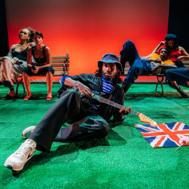 Dev Hynes Striking Pose With Guitar In Vibrant Setting Wallpaper