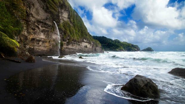 A black sand beach with a waterfall and lush green cliffs under a cloudy sky in Dominica, HD Desktop Wallpaper