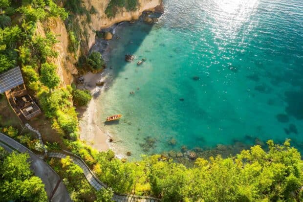 A bright aerial view of a coastal bay in Dominica showing clear turquoise water, sandy beach, and lush green cliffs, 2K Desktop Wallpaper