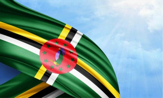 The image features the Dominica national flag waving against a bright blue sky with sunlight filtering through the clouds, 2K Desktop Wallpaper