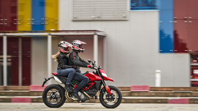 Ducati Hypermotard 950 Red Motorcycle Riding with Two Riders on Urban Street Background