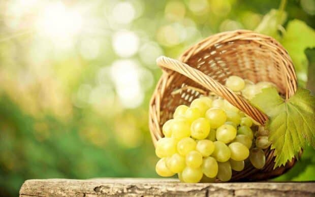 A bunch of fresh green grapes lies in a wicker basket on a wooden surface with a blurred bright green background, 2K Desktop Wallpaper
