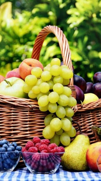 Fresh green grapes hang over a wicker basket filled with peaches, apples, and plums on a checkered cloth, HD Mobile Wallpaper