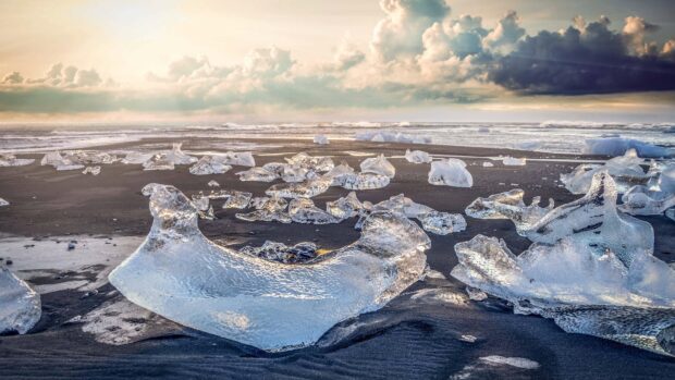 The Arctic displays glistening ice pieces scattered on dark sand near the shore, highlighting the cold climate and natural beauty