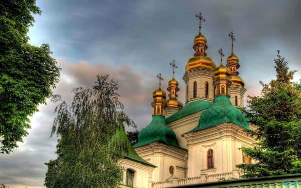 The golden domes of a historic church stand tall above lush green trees under a cloudy sky in Ukraine, 2K Desktop Wallpaper