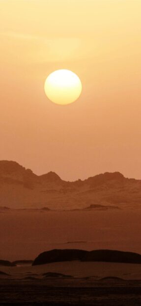 The golden sun sets behind the rocky desert hills in Algeria creating a warm peaceful scene, 2K Phone Wallpaper