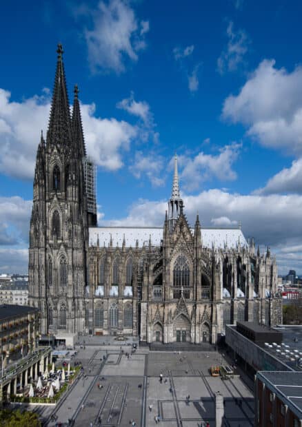 A Gothic cathedral with tall spires stands against a partly cloudy blue sky in a busy square, 2K Desktop Wallpaper
