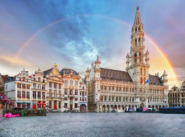 The Grand Place in Brussels is illuminated by a vibrant rainbow arching over historic buildings under a cloudy sky, featuring Brussels wallpapers, 2K Desktop W