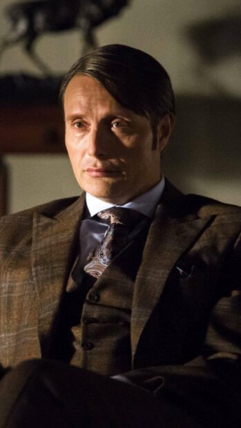 Hannibal Mads Mikkelsen sits thoughtfully in a dimly lit room wearing a patterned tie, 2K Phone Wallpaper