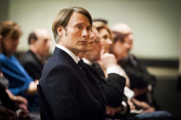 Hannibal Mads Mikkelsen sits attentively in a dark suit among other people in a formal setting, 2K Desktop Wallpaper