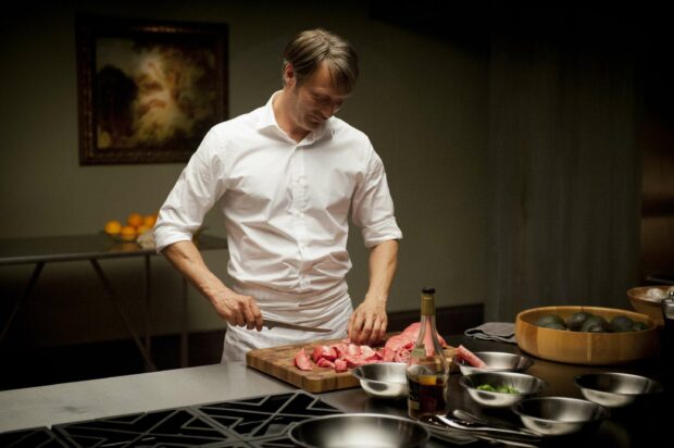 Hannibal Mads Mikkelsen carefully slicing meat on a wooden board in a dimly lit kitchen, 2K Desktop Wallpaper