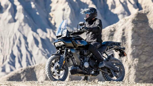 A rider on a Harley Davidson Pan America 1250 motorcycle is parked on rugged terrain with rocky mountains in the background, HD Desktop Wal