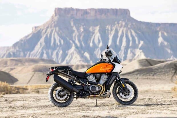 A Harley Davidson Pan America 1250 adventure motorcycle stands on a desert terrain with rocky mountains in the background