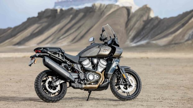 A Harley Davidson Pan America 1250 motorcycle parked on a sandy terrain with rocky hills in the background