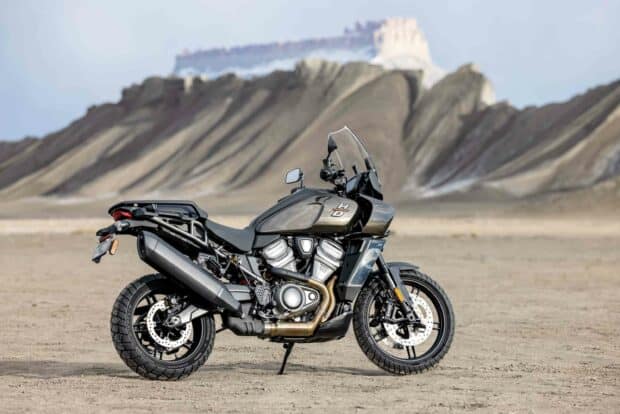 A Harley Davidson Pan America 1250 motorcycle is parked on a desert landscape with rocky hills in the background