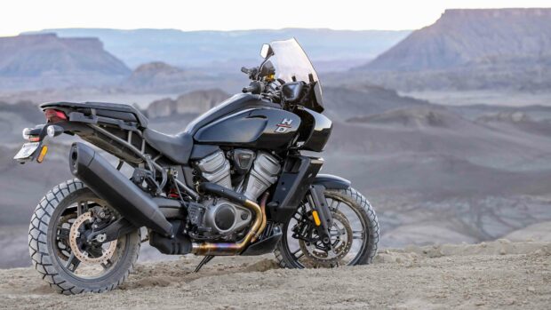 A Harley Davidson Pan America 1250 motorcycle is parked on sandy terrain with mountain views in the background