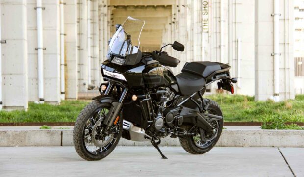 A black Harley Davidson Pan America 1250 motorcycle parked under a concrete bridge with green grass in the background