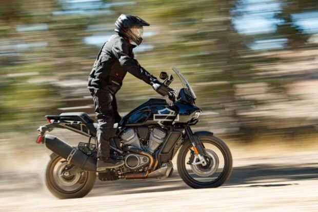 A rider wearing black gear is riding a Harley Davidson Pan America 1250 motorcycle at high speed on a dirt road, 2K Desktop Wallp