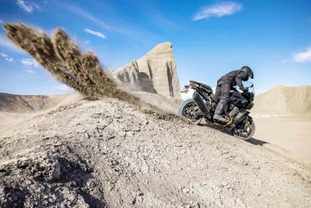 A rider on a Harley Davidson Pan America 1250 kicking up dirt on a desert trail under a clear sky, HD Desktop Wallpaper