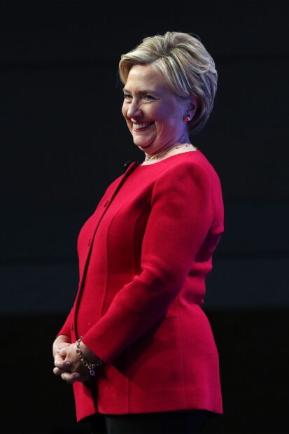 Hillary Clinton smiles warmly wearing a red jacket and standing against a dark background, 2K Desktop Wallpaper