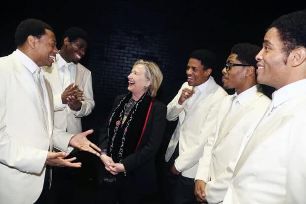 Hillary Clinton meets and laughs with a group of men dressed in white suits on a black stage, 2K Desktop Wallpaper