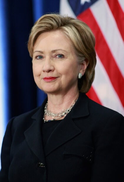 Hillary Clinton portrait standing confidently in front of the American flag wearing a black suit, 2K Mobile Wallpaper