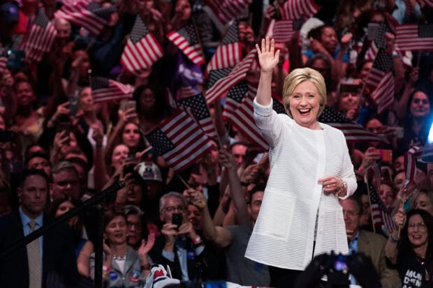 Hillary Clinton smiling and waving at a crowd holding American flags during a public event, 2K Desktop Wallpaper