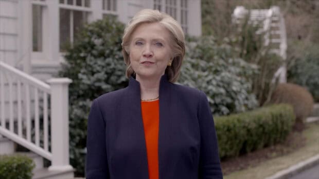 Hillary Clinton is standing outside in a dark jacket and red top near greenery, HD Desktop Wallpaper