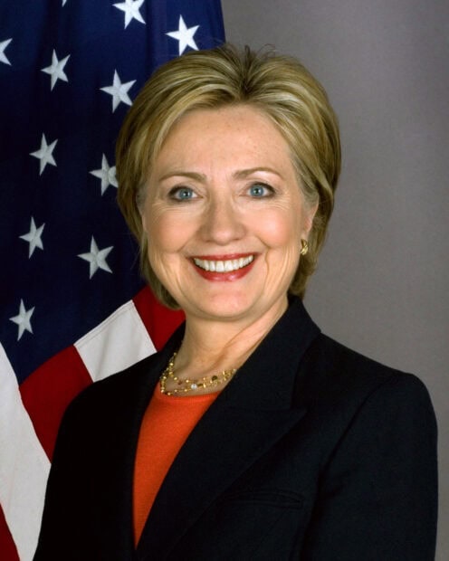 A smiling Hillary Clinton stands in front of the American flag wearing a black jacket, 2K Mobile Wallpaper