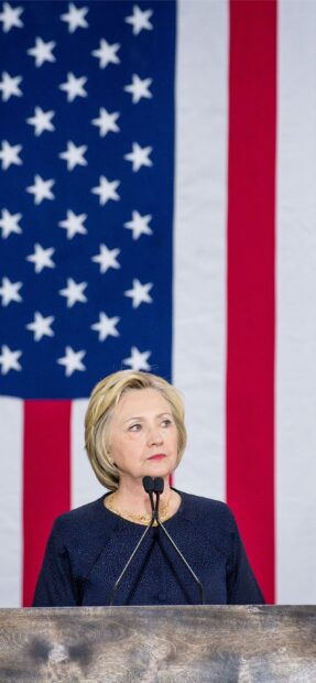 Hillary Clinton speaking at a podium with the American flag behind her, 2K Phone Wallpaper