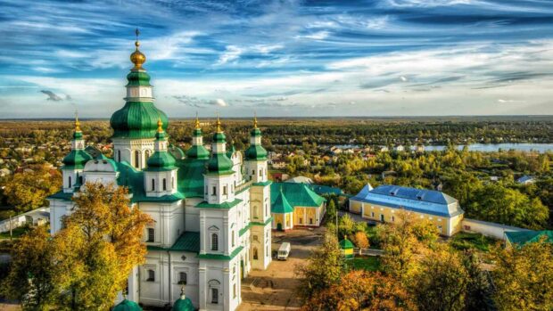 A scenic view of a historic Ukrainian church with green domes surrounded by autumn trees and a distant river, HD Desktop Wallpaper