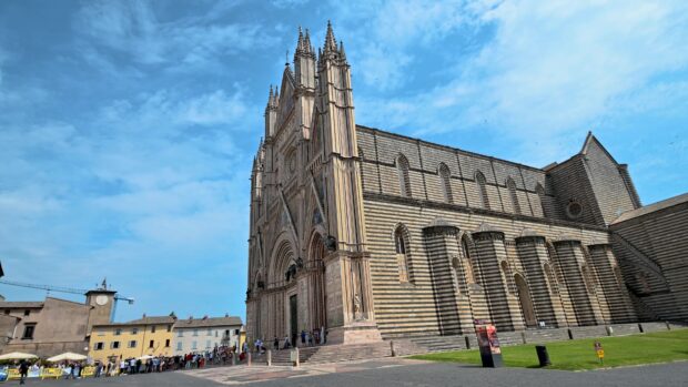 A historic cathedral with a unique striped stone facade and people gathered outside under a clear blue sky, 4K Desktop Wallpaper
