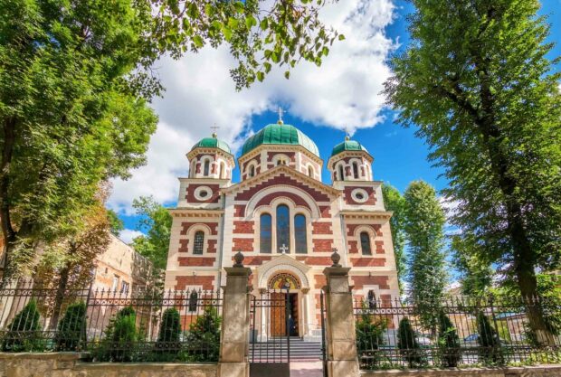 A historic church with distinctive green domes stands surrounded by lush trees under a partly cloudy blue sky in Ukraine, 2K Desktop Wallpaper
