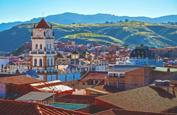 The image shows a historic clock tower and surrounding buildings with mountain hills in the background in Bolivia, HD Desktop Wallpaper