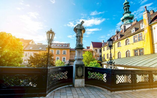 A historic statue stands on a small bridge with colorful old buildings and a bright blue sky in Bratislava, HD Desktop Wallpaper