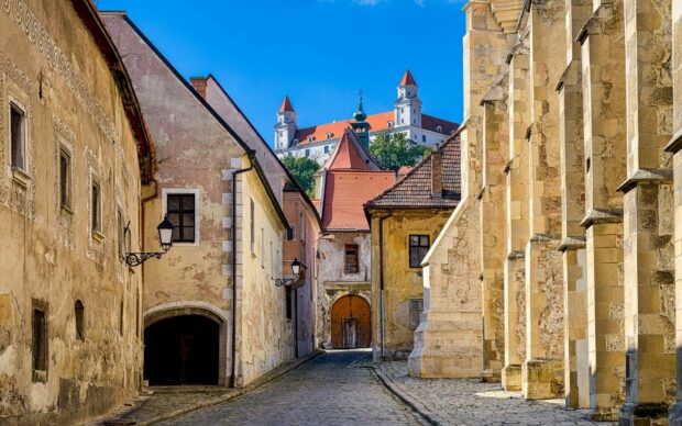 A cobblestone street leads through old buildings towards Bratislava Castle under a clear blue sky, HD Desktop Wallpaper