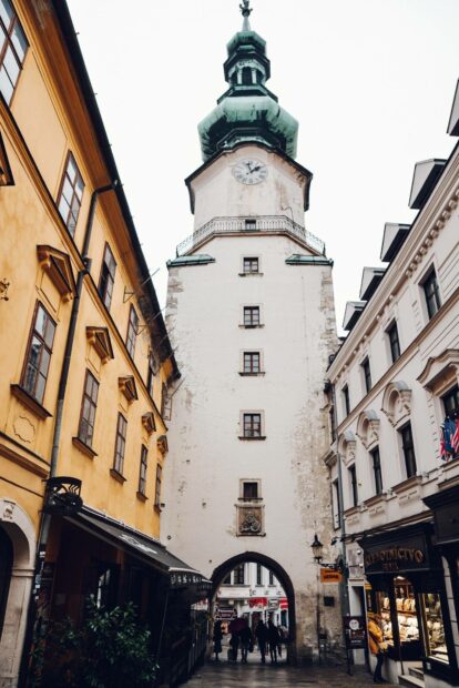 A historic tower stands among buildings in the center of Bratislava, featuring people walking beneath its archway, Bratislava Wallpapers, HD Mobile Wallp