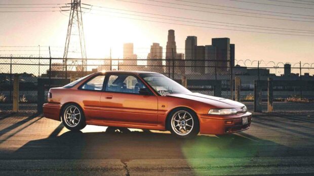 A red Honda Integra is parked by a fence with city buildings in the background at sunset, HD Desktop Wallpaper