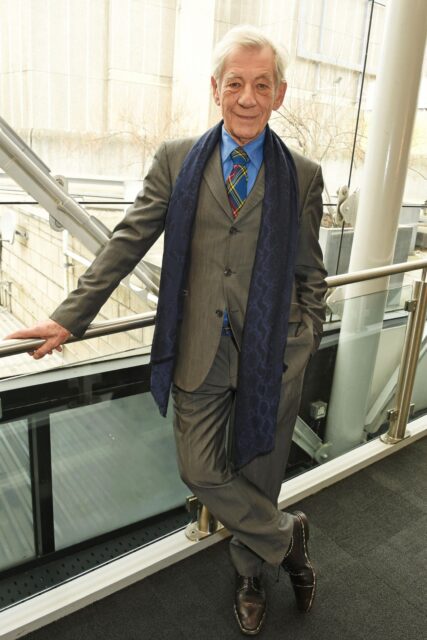Ian McKellen Stylish Pose In Grey Suit And Blue Scarf Wallpaper